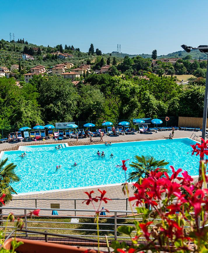 Piscina azzurra tra colline verdi e cielo sereno, fiori colorati in primo piano.