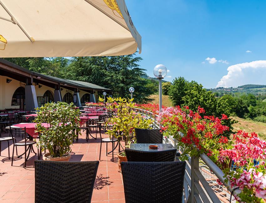 Terrazza soleggiata con tavoli rossi, fiori colorati e vista su colline verdi.